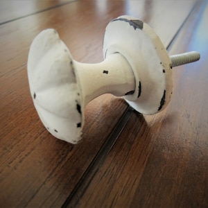 Może przedstawiać: Biały ceramiczny gałka z wytartym wykończeniem. Gałka ma okrągłą podstawę i żłobiony wierzchołek. Jest przymocowana do metalowego śruby.