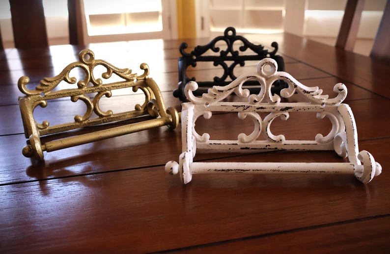 Puede incluir: Tres portarrollos de papel higi&eacute;nico decorativos en dorado, negro y blanco. Cada soporte presenta un dise&ntilde;o ornamental de volutas y una barra horizontal para sujetar el rollo de papel higi&eacute;nico. Los soportes son de metal y tienen un acabado envejecido.