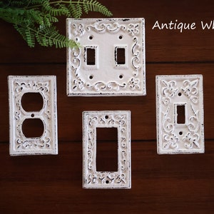 May include: Four white antique-style light switch and outlet covers with ornate designs. The covers are on a dark wood background. Antique White.
