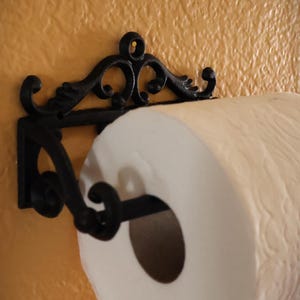 Puede incluir: Un portarrollos de papel de cocina de hierro fundido negro y ornamentado, montado en una pared texturizada de color naranja claro. El soporte tiene volutas decorativas y sostiene un rollo de papel de cocina blanco.