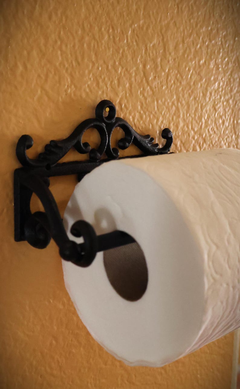 Puede incluir: Un portarrollos de papel de cocina de hierro fundido negro y ornamentado, montado en una pared texturizada de color naranja claro. El soporte tiene volutas decorativas y sostiene un rollo de papel de cocina blanco.