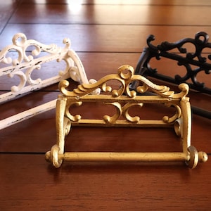 Puede incluir: Tres portarrollos de papel higi&eacute;nico decorativos en blanco, dorado y negro. Cada soporte tiene un dise&ntilde;o ornamental con volutas y una barra horizontal para el rollo. Los soportes son de metal y est&aacute;n sobre una superficie de madera.