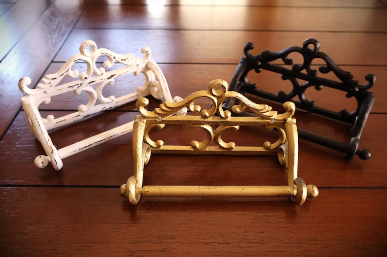 Puede incluir: Tres portarrollos de papel higi&eacute;nico decorativos en blanco, dorado y negro. Cada soporte tiene un dise&ntilde;o ornamental con volutas y una barra horizontal para el rollo. Los soportes son de metal y est&aacute;n sobre una superficie de madera.