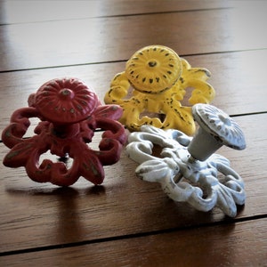 Op de afbeelding: Drie decoratieve kastknoppen met sierlijke bloemmotieven. De knoppen zijn rood, geel en lichtblauw geverfd.