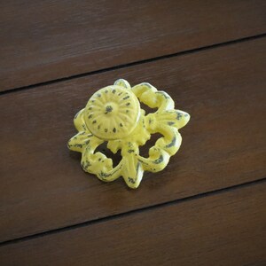 May include: A yellow metal drawer pull with a floral design. The pull has a distressed finish and is in the shape of a flower.
