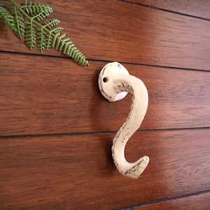 Puede incluir: Gancho de hierro fundido blanco con un diseño decorativo de espiral. El gancho está montado en una pared de madera.