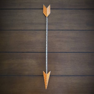 May include: A simple orange arrow with a white string hanging from it. The arrow is pointing down and is against a dark brown wood background.