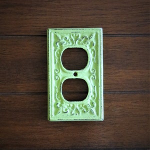May include: A light green outlet cover with an ornate design. The cover is made of metal and has a distressed finish.