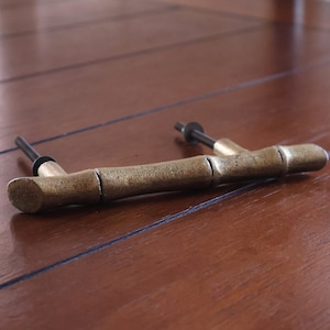 May include: A bronze-colored cabinet pull with a bamboo design. The handle is horizontal, with a series of segmented sections mimicking bamboo stalks. The mounting hardware is black. The pull is set against a dark wood surface.
