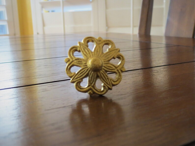 May include: A gold-coloured metal decorative knob with a floral design. The knob is circular and features intricate details.