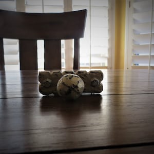Op de afbeelding: Een witte gietijzeren deurknop met een verweerde afwerking. De knop ligt op een houten tafel met een stoel op de achtergrond.