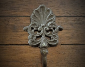 May include: An ornate, antique-style metal hook with a decorative, scrolling design. The hook is a dark bronze colour and is mounted on a wooden surface. The hook is designed for hanging items.