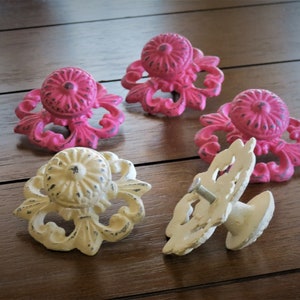 Könnte beinhalten: Fünf verzierte Schrankknöpfe, vier sind rosa und einer ist cremefarben. Die Knöpfe haben ein florales Design und sind aus Metall.