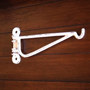 Op de afbeelding: Wit metalen wandrek met een haak aan het uiteinde. Het rek is gemonteerd op een donker houten oppervlak.