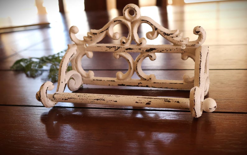 Puede incluir: Un portarrollos de papel higi&eacute;nico de metal blanco envejecido con adornos de volutas y una parte superior decorativa. El soporte tiene una barra para el rollo de papel higi&eacute;nico y est&aacute; dise&ntilde;ado para montarse en la pared. El acabado tiene un aspecto r&uacute;stico y vintage.