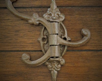 Puede incluir: Un gancho triple de hierro fundido rústico de estilo antiguo con un diseño decorativo y ornamentado. El gancho está montado en una superficie de madera.