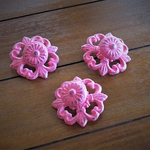 May include: Three pink metal drawer pulls with a floral design. The pulls have a distressed finish and are perfect for adding a touch of vintage charm to any home decor project.