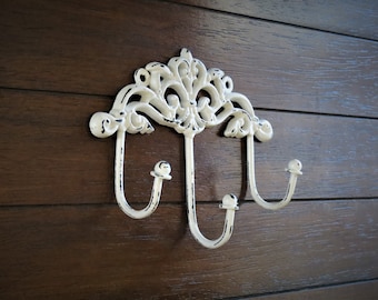 Puede incluir: Un gancho de pared de metal blanco con un diseño decorativo de desplazamiento y tres ganchos. El gancho está montado en una pared de madera marrón.