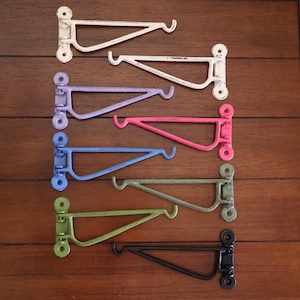 Może przedstawiać: Siedem metalowych wsporników do półek w różnych kolorach, w tym białym, lawendowym, różowym, niebieskim, zielonym i czarnym. Wsporniki mają ten sam rozmiar i kształt, z prostym, zakrzywionym wzorem.