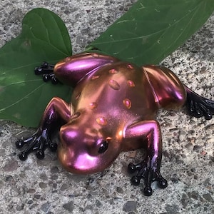 May include: A purple and gold frog figurine with black painted details. The frog is sitting on a green leaf.