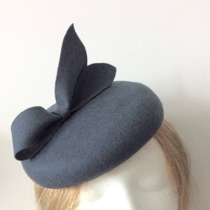 May include: A gray felt hat with a large bow on the side. The hat is a pillbox style and is perfect for a special occasion.