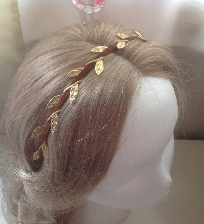 May include: A brown headband with gold leaf accents. The headband is worn on a mannequin with blonde hair.