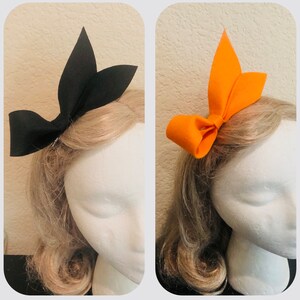 May include: Two felt hair bows, one black and one orange, are displayed on mannequin heads. The bows have a classic bow shape with pointed accents. The mannequin heads have blonde hair and are set against a neutral background.
