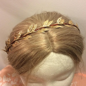 May include: A gold leaf crown headband with a brown wire base. The headband is worn on a mannequin with blonde hair.