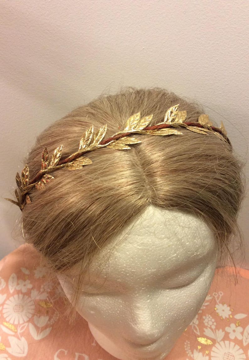 May include: A gold leaf crown headband with a brown wire base. The headband is worn on a mannequin with blonde hair.