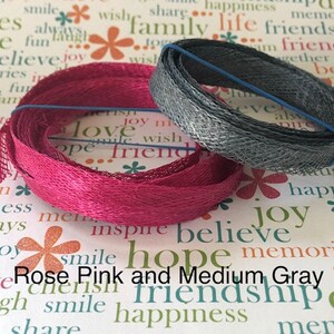 May include: Two spools of ribbon, one pink and one gray. The pink ribbon is a woven, textured fabric. The gray ribbon is a metallic mesh. The ribbons are on a white background with a pattern of flowers and words.