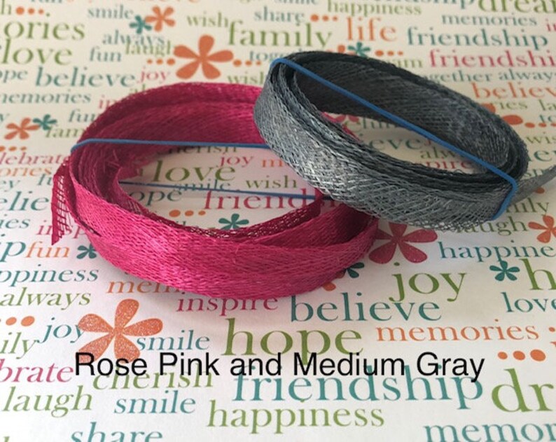 May include: Two spools of ribbon, one pink and one gray. The pink ribbon is a woven, textured fabric. The gray ribbon is a metallic mesh. The ribbons are on a white background with a pattern of flowers and words.