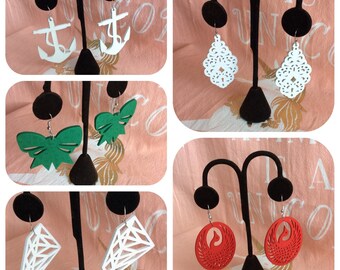 May include: A set of 10 pairs of wooden earrings displayed on a black stand. The earrings are in various shapes and colors, including white anchors, green bows, white geometric shapes, and red phoenixes.