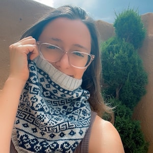 May include: A knitted cowl with a geometric pattern in navy blue and light gray. The cowl is worn around the neck, partially covering the face. The background includes greenery and a cloudy sky.