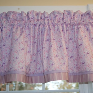 May include: A ruffled valance curtain with a floral pattern in shades of pink, purple, and white. The bottom edge features a striped border in coordinating colors. The curtain is hanging above a window.