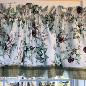 May include: A valance curtain with a white background, featuring a floral pattern of green leaves, white poinsettias, and brown pinecones. The bottom edge has a green plaid border. The curtain is gathered at the top.