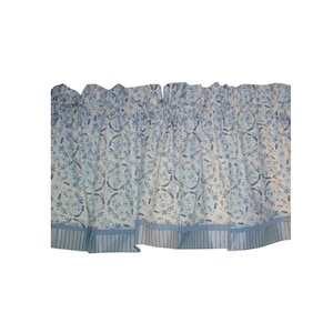May include: A window valance featuring a light blue and white floral pattern with wreaths and leaves. The fabric has a gathered top for rod insertion and a bottom border with vertical blue and white stripes, creating a pleated, layered appearance.