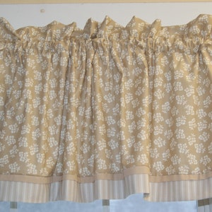 May include: A beige floral valance with white flower patterns. The valance has a ruffled top and a striped bottom border. The fabric is a light, neutral color, suitable for a variety of interior design styles.