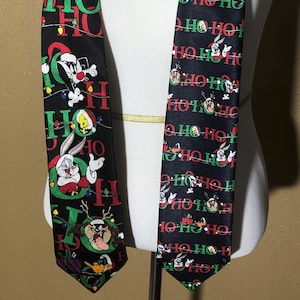 Peut inclure: Une cravate en soie noire sur le thème de Noël avec Bugs Bunny, Daffy Duck et Titi. La cravate a un motif répété de "Ho Ho Ho" en texte rouge et vert.