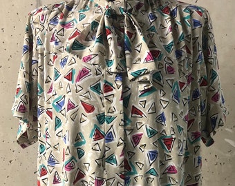 May include: A beige blouse with a short sleeve and a bow tie. The blouse has a geometric pattern of colorful triangles.