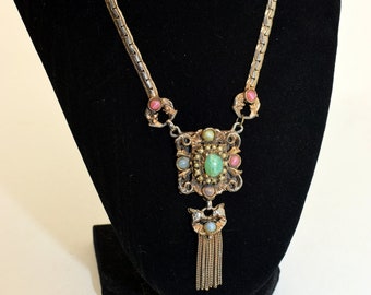 May include: A gold chain necklace with a green, pink and blue stone pendant. The pendant is a square with a tassel hanging from the bottom.