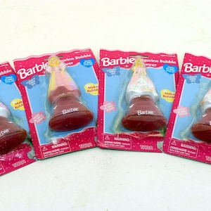 Puede incluir: Cuatro juguetes de soplador de burbujas de figuras de Barbie en su embalaje original. Cada paquete presenta un diseño rosa y azul con el logotipo de Barbie y una figura de muñeca. Los sopladores de burbujas tienen una base roja y una muñeca encima.