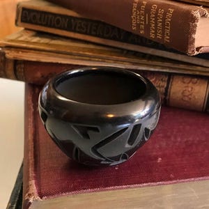 May include: A small, black ceramic bowl with carved geometric designs. The bowl is round with a wide opening and sits atop a stack of antique books with red and brown covers. The books have aged, worn pages.