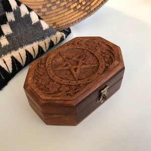 May include: A dark brown, wooden box with a pentagram carved on the lid. The box has a hexagonal shape and is decorated with floral and star patterns. A metal clasp secures the box.