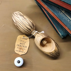 以下が含まれることがあります： 「A Genuine Walnut Nut Cracker」と書かれたタグが付いた、木製のヴィンテージのクルミ割り器。クルミ割り器は薄茶色で、彫刻が施されています。白と黒のノブがクルミ割り器の隣にあります。