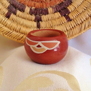 Puede incluir: Un pequeño recipiente redondo de madera roja con un diseño pintado en blanco, naranja y beige. El recipiente se encuentra sobre una superficie blanca con una cesta tejida en el fondo. La cesta tiene un diseño circular con colores marrón, naranja y beige.