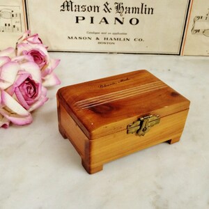 May include: A small, rectangular wooden box with a hinged lid and brass clasp. The box is a warm, reddish-brown color and has a simple, elegant design. The text "Glennie, Mich." is inscribed on the lid. A bouquet of pink roses is in the background.