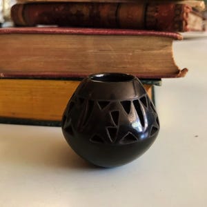 Könnte beinhalten: Eine kleine, schwarze Keramikvase mit rundem Körper und geometrischen Ausschnitten. Die Vase wird vor einem Stapel alter Bücher mit abgenutzten Einbänden präsentiert, was eine Vintage-Ästhetik erzeugt.