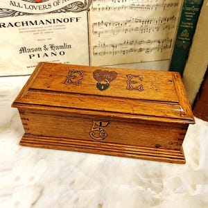 May include: A rectangular wooden box with a hinged lid. The box is a warm brown colour and features decorative carvings, including the letter "E" and a heart-shaped lock. The box is likely vintage and sits on a marble surface.