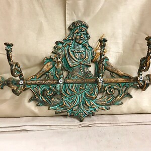 May include: A decorative wall-mounted coat rack with a green and gold finish. The rack features a central figure of a woman with flowing hair and a flowing gown. The rack has three hooks for hanging coats, hats, or other items.