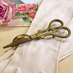 May include: Antique brass scissor-style wick trimmer. The tool features a scissor handle design with a curved blade and a small, rounded container. The trimmer is set against a backdrop of a white cloth, a greeting card, and a pink rose.